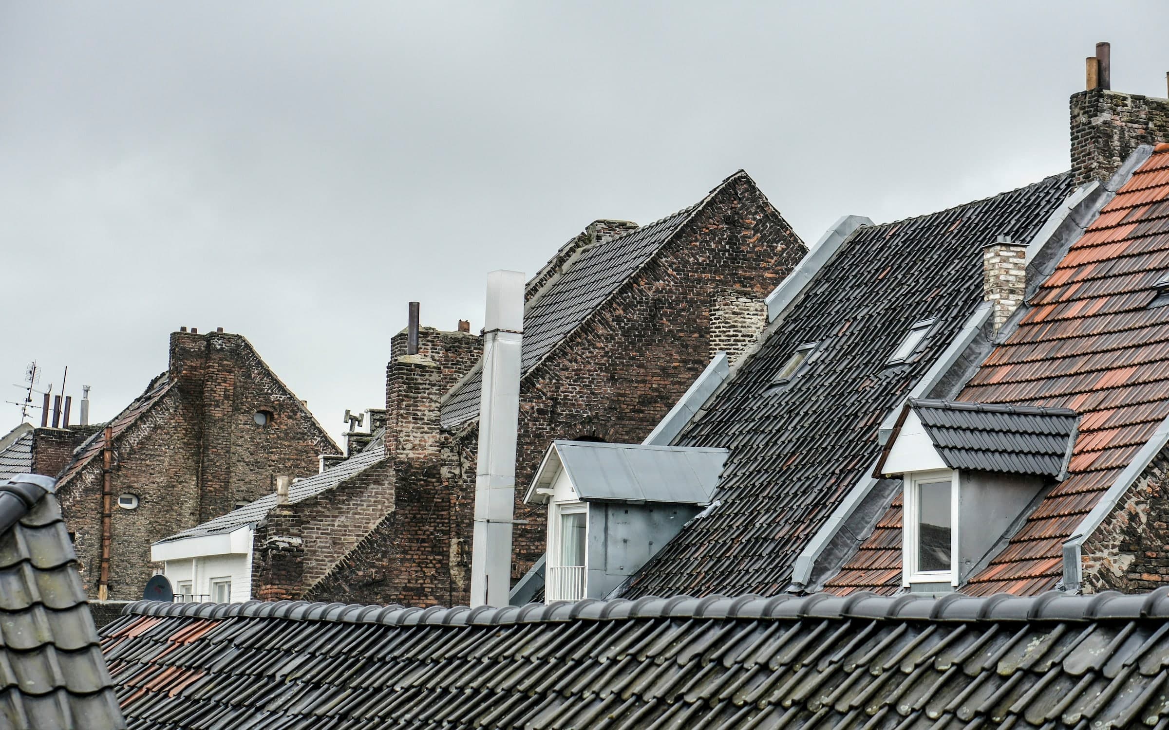 Toits anciens en tuiles anthracite et terre cuite, lucarnes habillées de zinc et cheminées en brique, sous un ciel gris